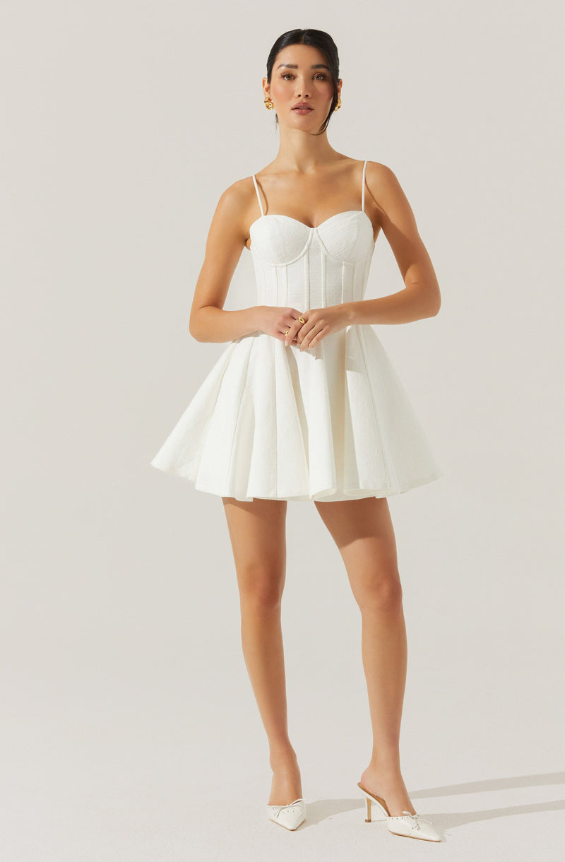 A woman is wearing a white dress with a fitted bodice featuring vertical seams and a sweetheart neckline. The dress has thin spaghetti straps and a full, flared skirt with pleats. She is also wearing white high-heeled shoes with a decorative bow detail on the toe.