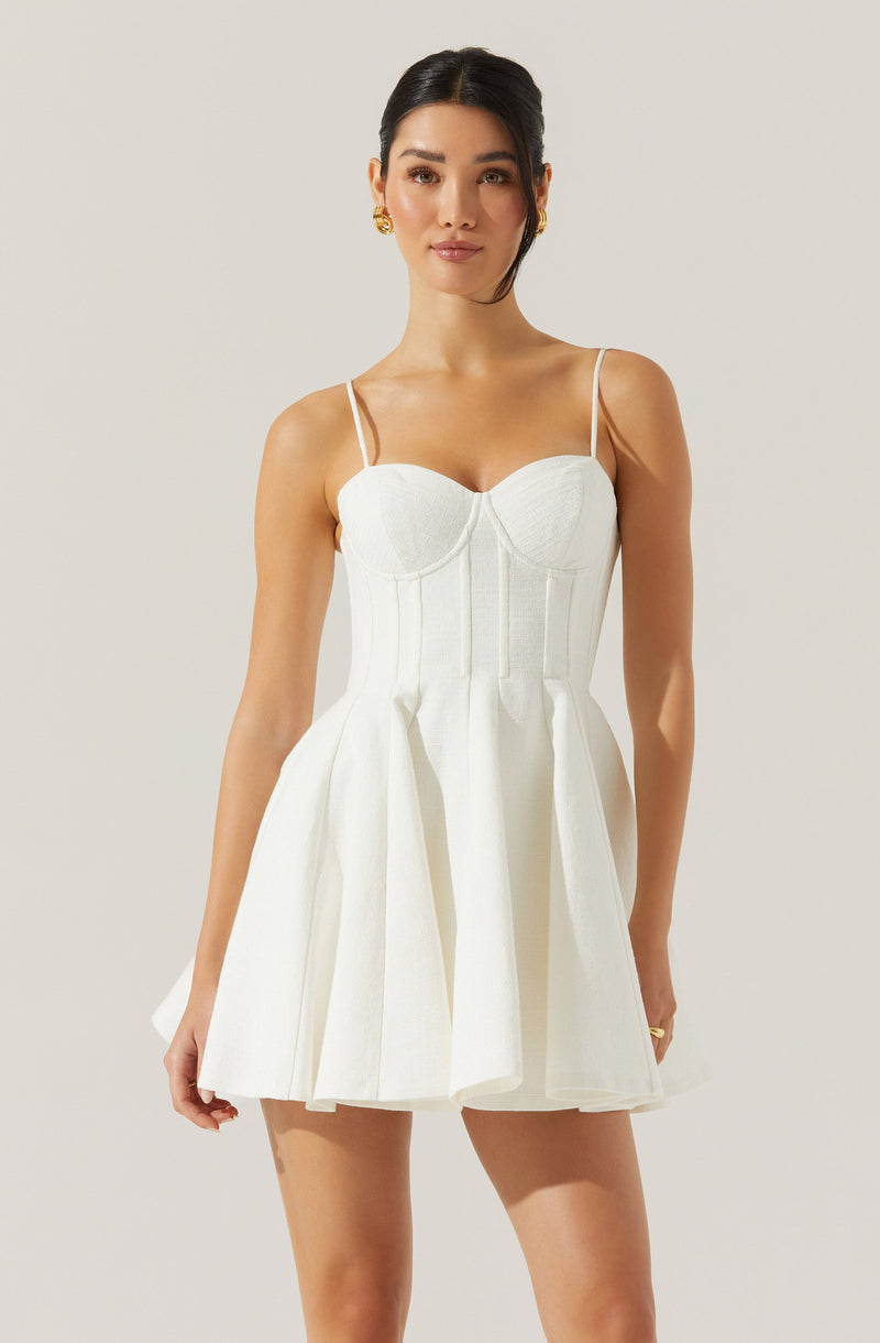 A woman is wearing a sleeveless, white dress with thin spaghetti straps. The dress features a fitted bodice with subtle vertical seam details and a flared, pleated skirt that reaches mid-thigh.