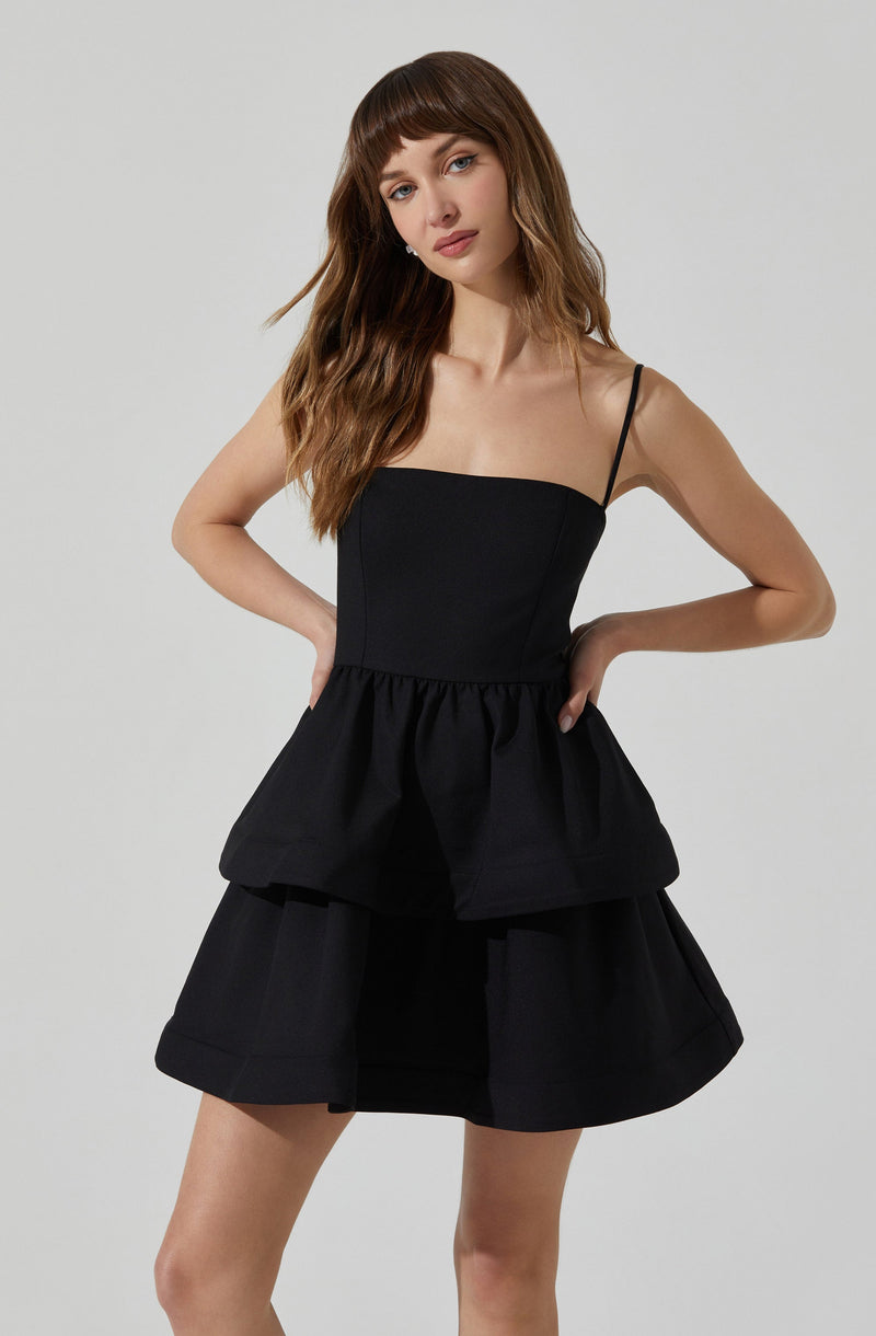 A young woman is wearing a black sleeveless dress with thin spaghetti straps. The dress has a fitted bodice and a gathered peplum waist. The skirt portion of the dress is layered with a wide, flared design.