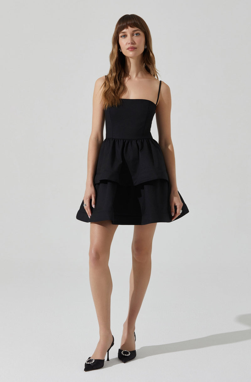 A woman is wearing a black dress with a fitted bodice and thin spaghetti straps. The dress features a peplum-style overlay at the waist and a flared skirt. She is also wearing black high-heeled shoes with decorative embellishments on the toes.
