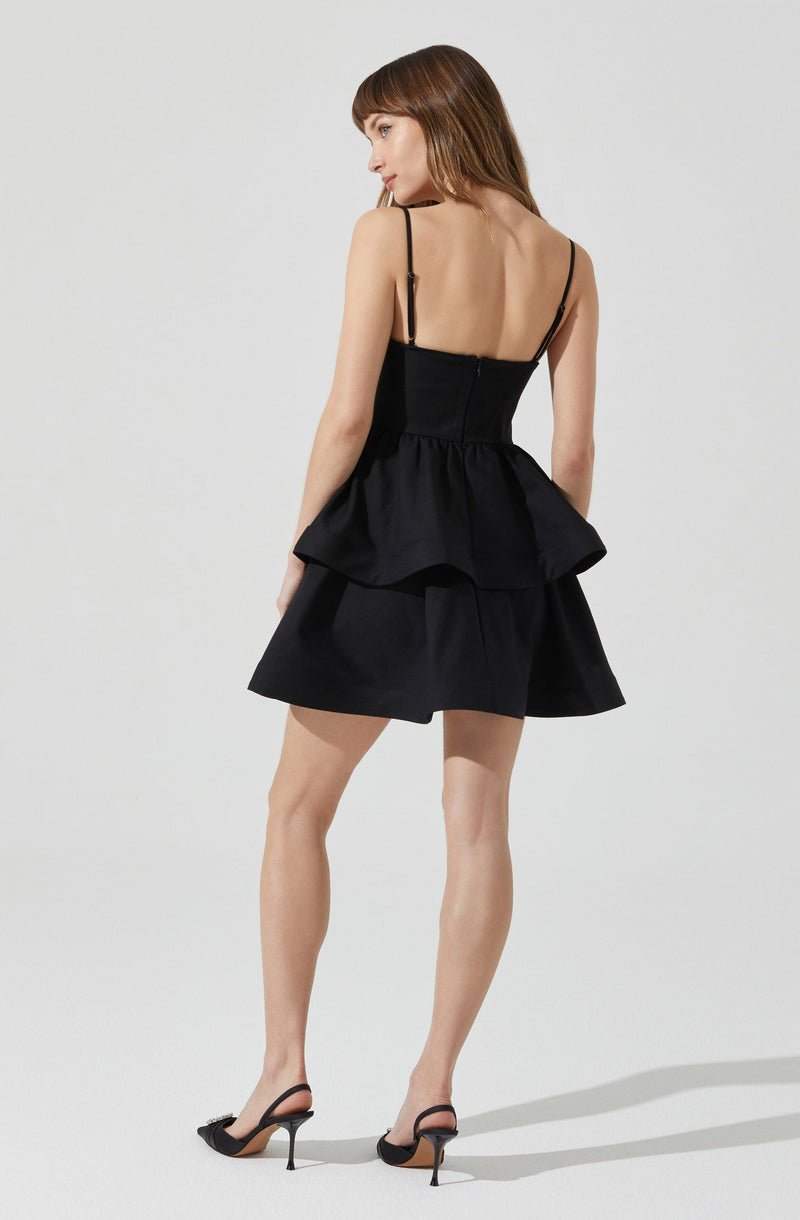 A woman is wearing a black sleeveless dress with thin spaghetti straps. The dress features a fitted bodice and a ruffled peplum waist that extends into a layered, flared skirt. She is also wearing black high-heeled shoes with ankle straps.