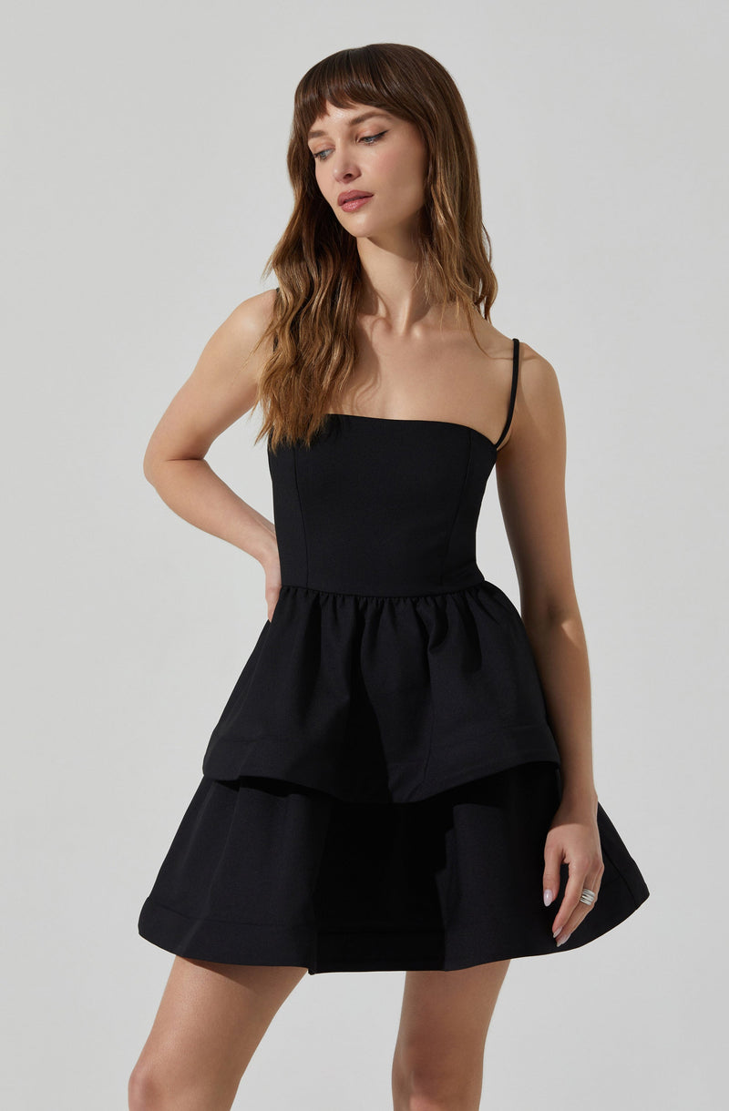A woman is wearing a black sleeveless top with thin spaghetti straps and a peplum waist. She pairs it with a black skirt that has a layered, flared design.