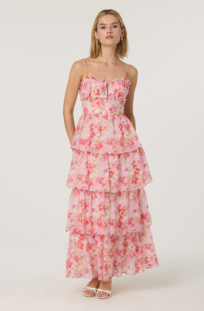 A woman is wearing a sleeveless, tiered maxi dress with thin spaghetti straps. The dress features a ruffled bodice with a zipper in the front, and it has a pink floral pattern with shades of pink, peach, and coral on a sheer fabric. The dress has multiple layered sections, creating a flowing, bohemian look. She is also wearing light-colored open-toe sandals.