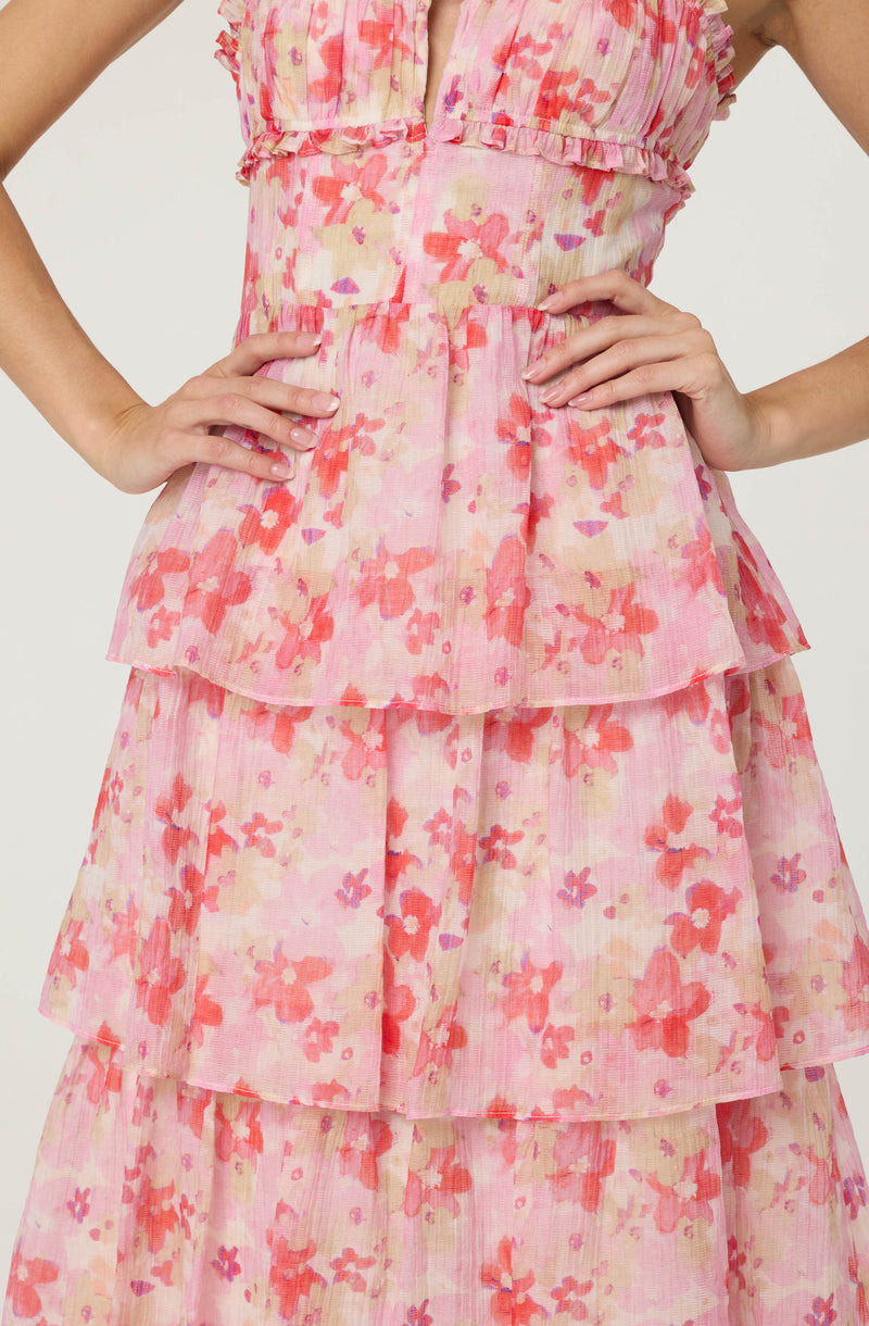 A pastel pink dress with a floral pattern featuring shades of coral, lavender, and beige. The dress has a sleeveless design with ruffle details along the armholes and multiple tiered, ruffled layers on the skirt, creating a flowy and layered look. The fabric appears lightweight and slightly sheer.