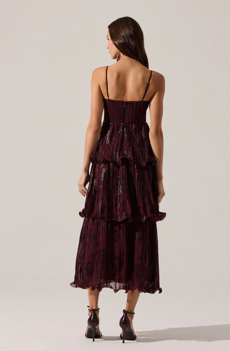 A woman is wearing a sleeveless, full-length dress with thin spaghetti straps. The dress is deep burgundy with a shiny, textured pattern, featuring multiple layered, ruffled tiers that add volume and movement. The fabric appears to be lightweight and semi-transparent with a delicate, slightly crinkled appearance. She pairs the dress with dark high-heeled shoes with ankle straps.