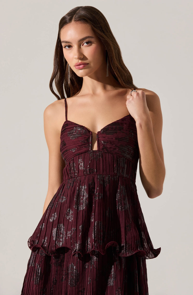 A woman is wearing a spaghetti strap dress in a deep burgundy color with a shiny, metallic black pattern. The dress features a fitted bodice with vertical and horizontal pleats, a small front keyhole detail, and a layered, ruffled skirt with a wavy hemline.