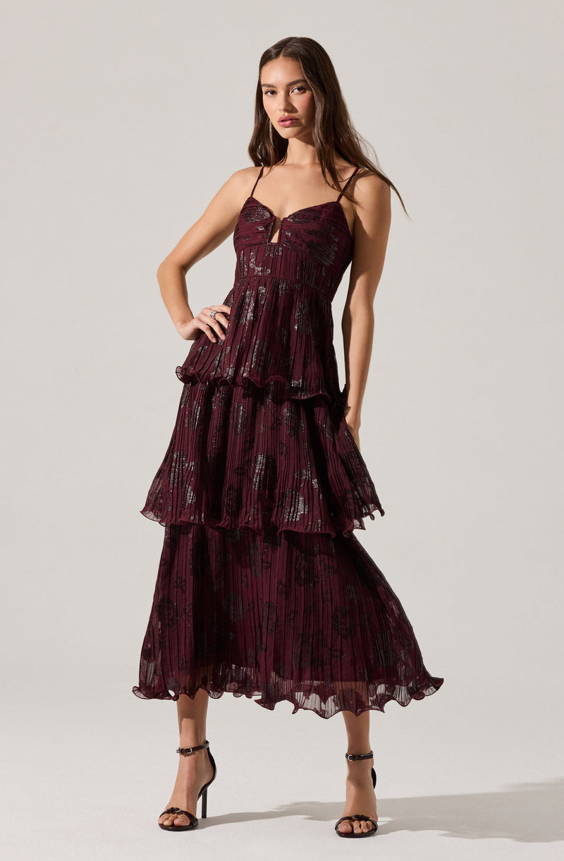 A woman is wearing a deep burgundy-colored dress with thin spaghetti straps. The dress features a fitted bodice with a sweetheart neckline and a small vertical slit at the center. The skirt has multiple layered, ruffled tiers made from a sheer, lightweight fabric with a subtle floral and striped pattern in black. She is also wearing black high-heeled sandals with ankle straps.