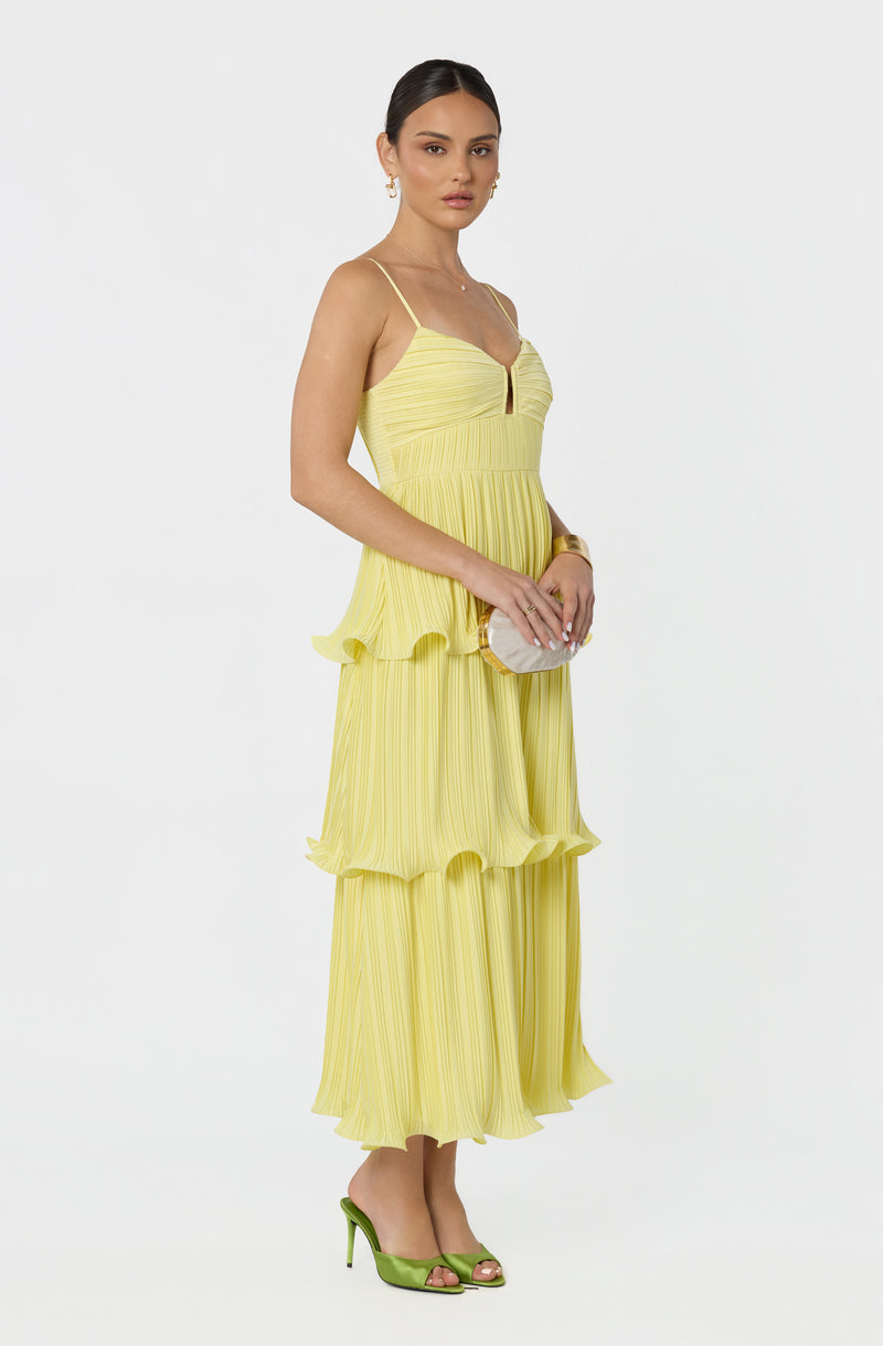 Woman in yellow pleated maxi dress with spaghetti straps, holding a white clutch, wearing green heels.