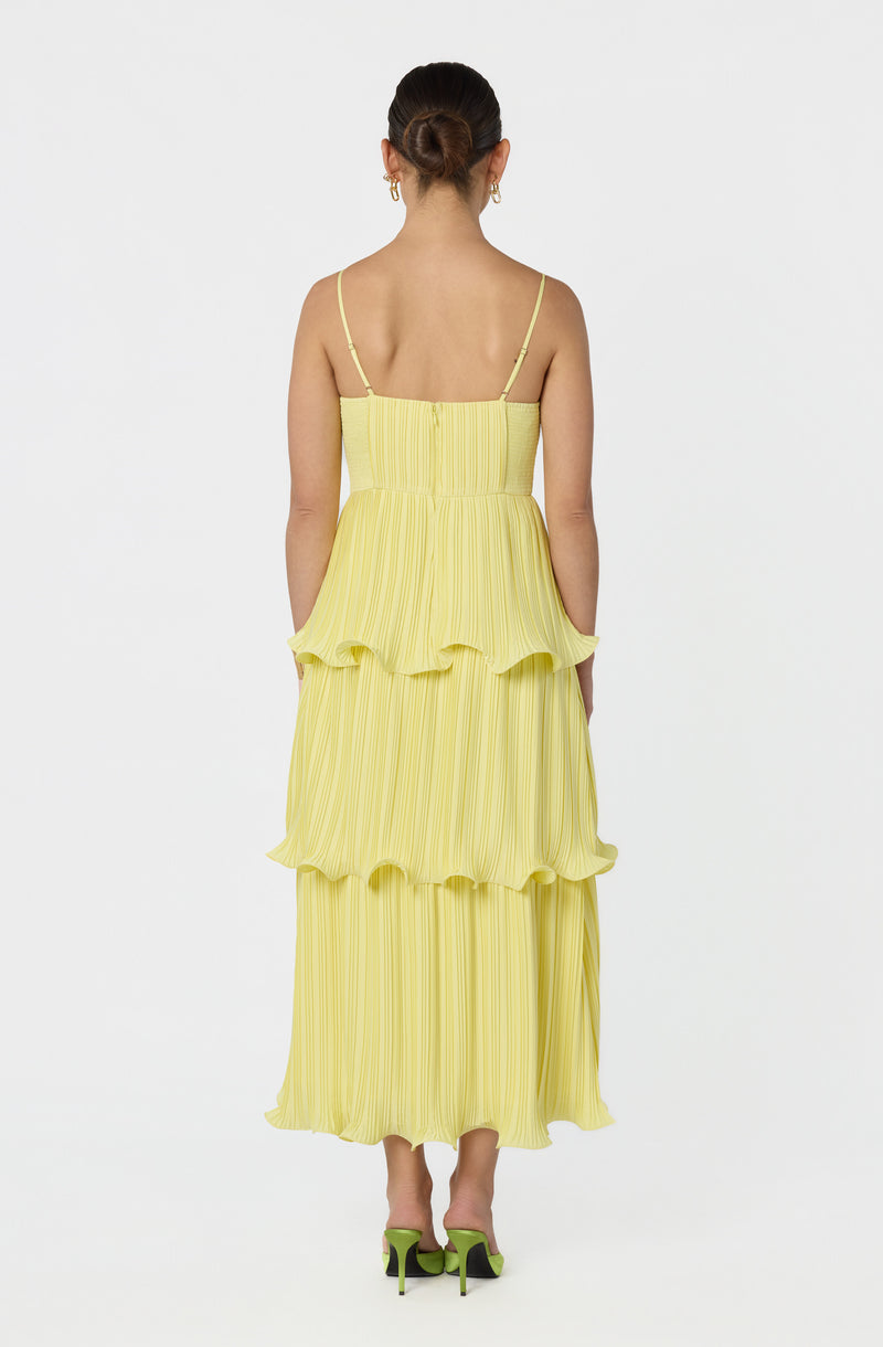 Woman in a yellow, pleated, tiered summer dress with thin spaghetti straps, wearing green high-heeled shoes.
