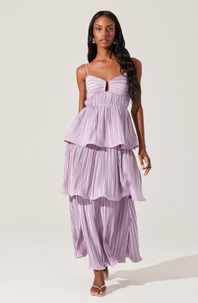 A woman is wearing a lavender-colored dress with thin spaghetti straps. The dress features a fitted bodice with a small keyhole cutout in the center. The skirt has three layered, pleated tiers, each with a slightly ruffled edge, creating a flowing and tiered look. The fabric is lightweight and has vertical pinstripes throughout. She is also wearing open-toe high-heeled sandals.