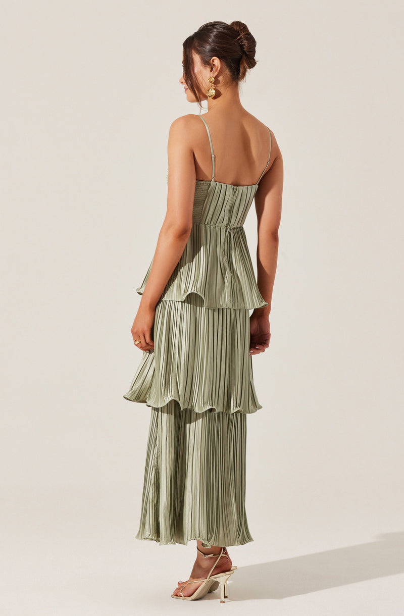 A woman is wearing a light green, pleated, strap-shoulder dress with thin spaghetti straps. The dress features a fitted bodice and a layered, ruffled skirt with vertical stripes. She has paired the dress with nude high-heeled sandals and gold earrings.