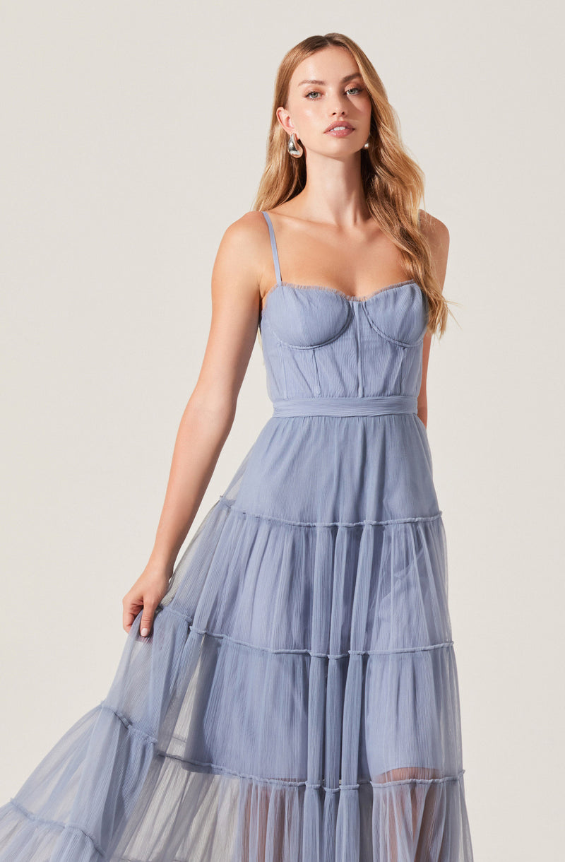 The woman is wearing a light blue, sleeveless dress with thin spaghetti straps. The bodice features a sweetheart neckline with ruched detailing. The waistline is fitted, and the skirt is long and tiered with horizontal seams and sheer fabric panels, creating a layered, flowing look. The dress has a soft, airy appearance with a subtle gathered texture throughout.