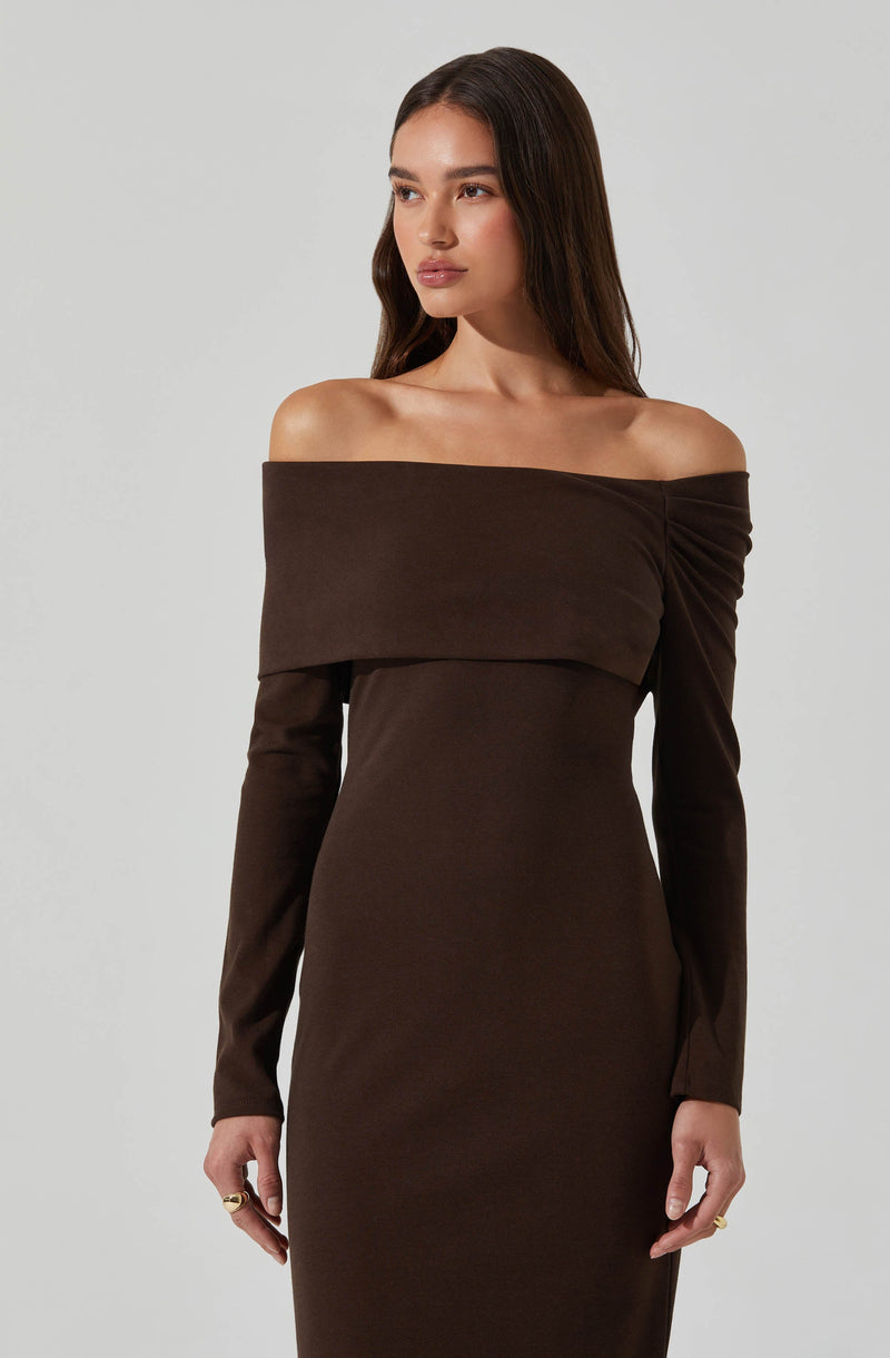 A woman is wearing a fitted, off-the-shoulder dress in dark brown fabric. The dress features a wide, folded over neckline that drapes across the shoulders and upper arms, with long sleeves extending to the wrists. The material appears smooth and slightly stretchy, accentuating the figure.