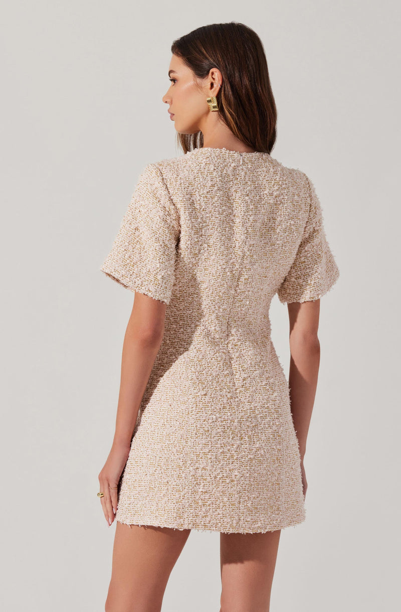 A woman is wearing a short, fitted dress made of a textured, woven fabric in a light beige or cream color. The dress has short sleeves and a high neckline, and it extends to mid-thigh length. She is accessorized with bold gold earrings and a gold ring on her right hand.