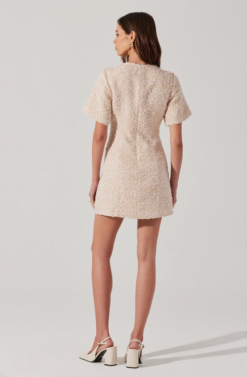 A woman is wearing a short, textured beige dress with short sleeves. The dress has a loose fit and a subtle seam running vertically down the back. She is also wearing white high-heeled sandals with an ankle strap and gold earrings.