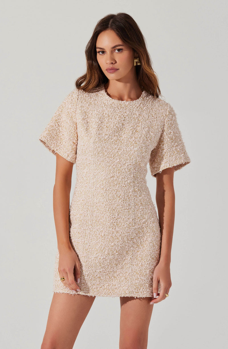 A woman is wearing a short, cream-colored textured dress with short, loose sleeves and a round neckline. The fabric appears woven with a multicolored pattern, giving it a slightly fuzzy appearance. She has gold earrings and rings, and her hair is styled in loose waves.
