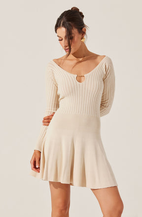 A woman is wearing a beige, knitted dress with a ribbed texture. The dress has a deep neckline with a small circular cutout detail at the center. The sleeves are long, and the skirt is pleated, giving it a slightly flared appearance.