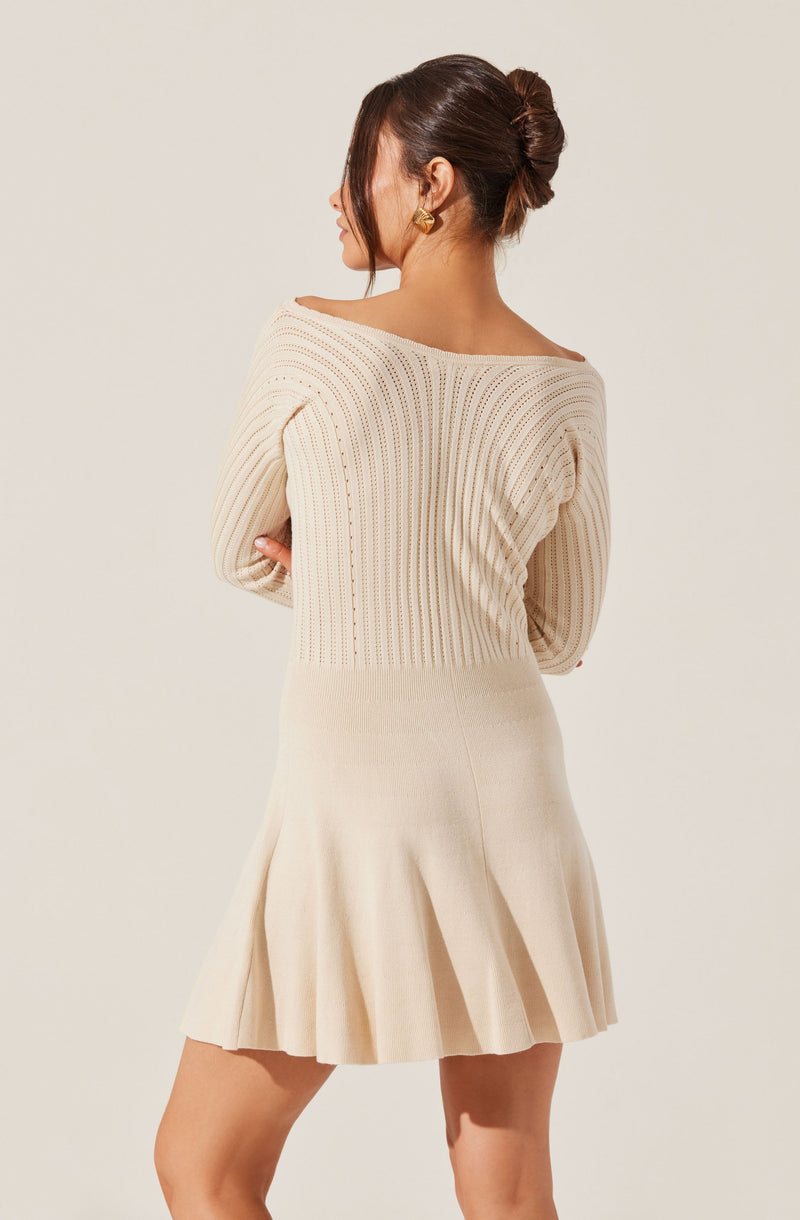 A woman is wearing a cream-colored knitted sweater with a wide, rounded neckline and long sleeves. She is also wearing a matching cream-colored skirt with pleats that fall above the knees. She has her hair styled in an elegant bun and is accessorized with gold earrings.
