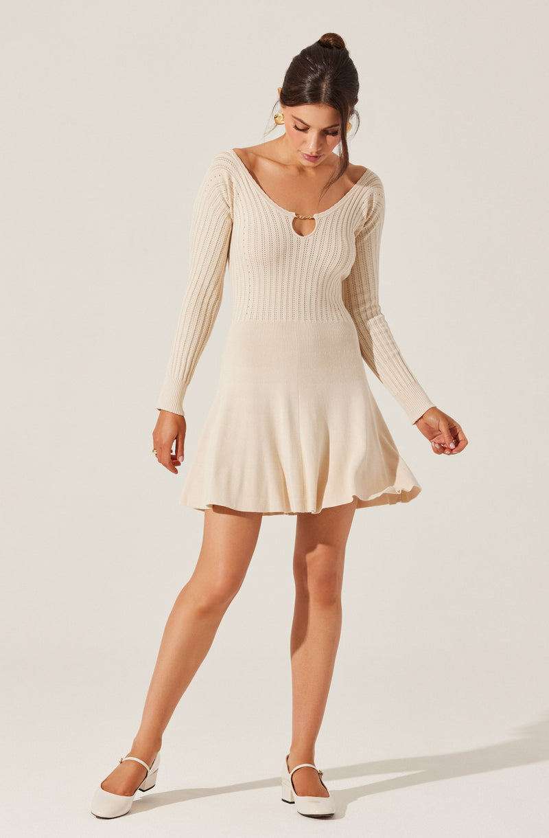 A woman wearing a cream-colored, long-sleeved knit dress with a ribbed texture. The dress has a keyhole opening at the neckline with a small metal ring detail. The skirt portion of the dress is flared and falls above the knees. She is also wearing white heels.