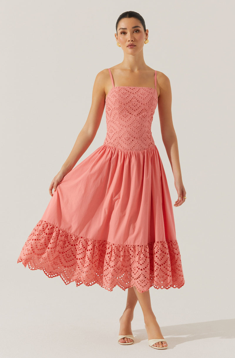 A woman is wearing a pink dress with thin shoulder straps. The top part of the dress is fitted and features an intricate, eyelet lace pattern. The skirt is full and gathered, made of a lightweight fabric, with a wide band of eyelet lace trim near the hem that matches the top's pattern. She is also wearing open-toed, high-heeled sandals.