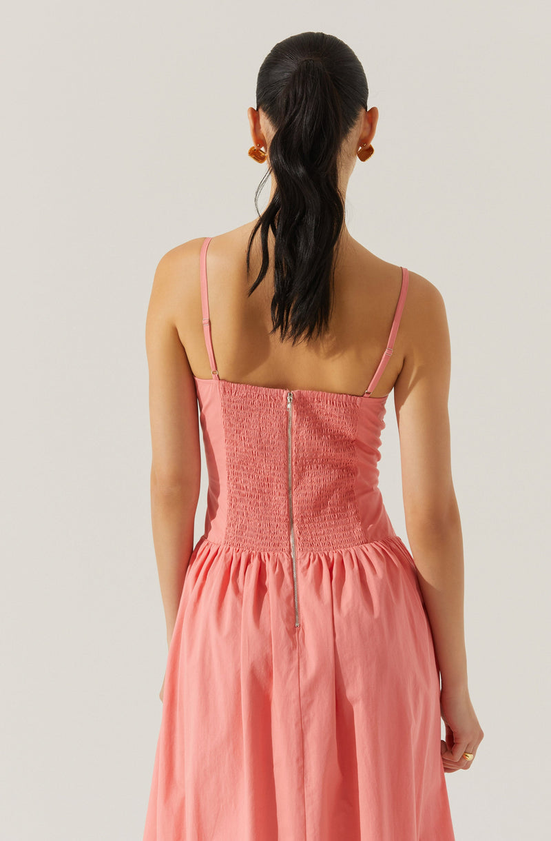 A woman is wearing a sleeveless, pink dress with thin spaghetti straps. The dress features a fitted bodice with smocking detail at the back and a vertical zipper running down the center. The skirt portion of the dress is gathered and flows loosely. The woman has dark hair styled in a low ponytail and is wearing large, square-shaped earrings.