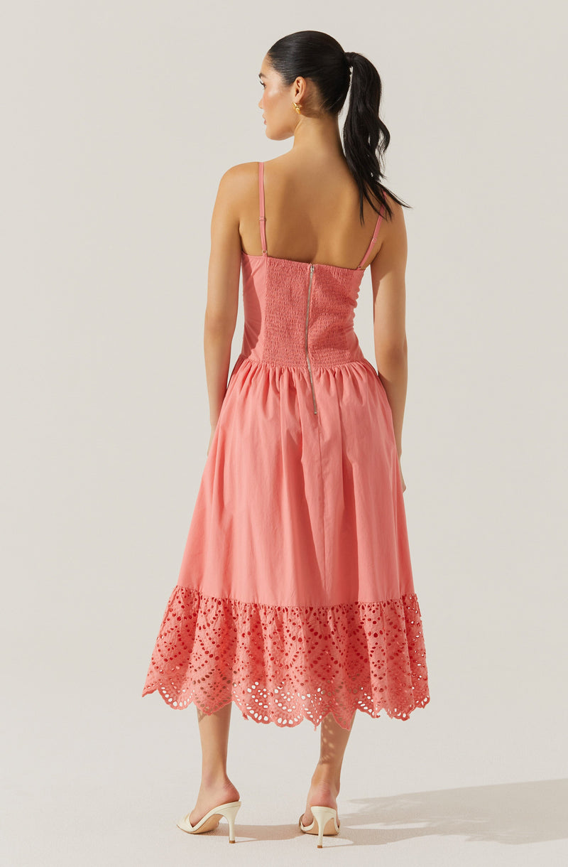 A woman is wearing a sleeveless, spaghetti strap pink dress. The dress features a fitted, smocked bodice with a zipper at the back, and a full skirt that falls below the knees. The hem of the skirt is decorated with a wide band of eyelet lace with a scalloped edge.