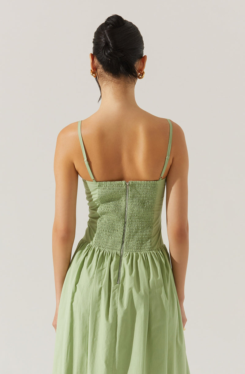 A woman is wearing a light green dress with thin spaghetti straps. The dress features a fitted, ruched bodice with a visible zipper down the center back and a gathered skirt starting at the waist.