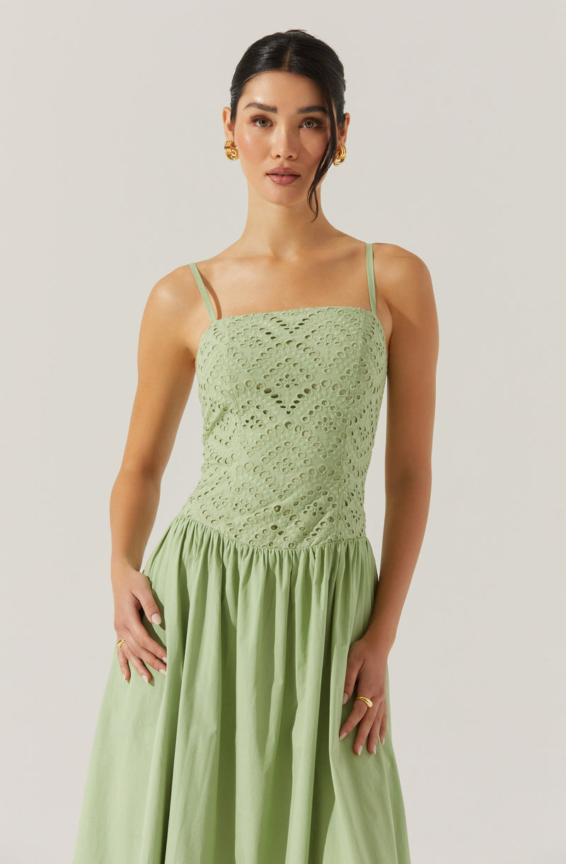 A woman is wearing a light green dress with thin spaghetti straps. The top portion of the dress features a textured, eyelet fabric with circular and diamond-shaped cutouts. The skirt of the dress is gathered at the waist, flowing freely with a smooth, solid fabric. She has dark hair styled in a sleek updo, and she is accessorizing with gold earrings and rings.