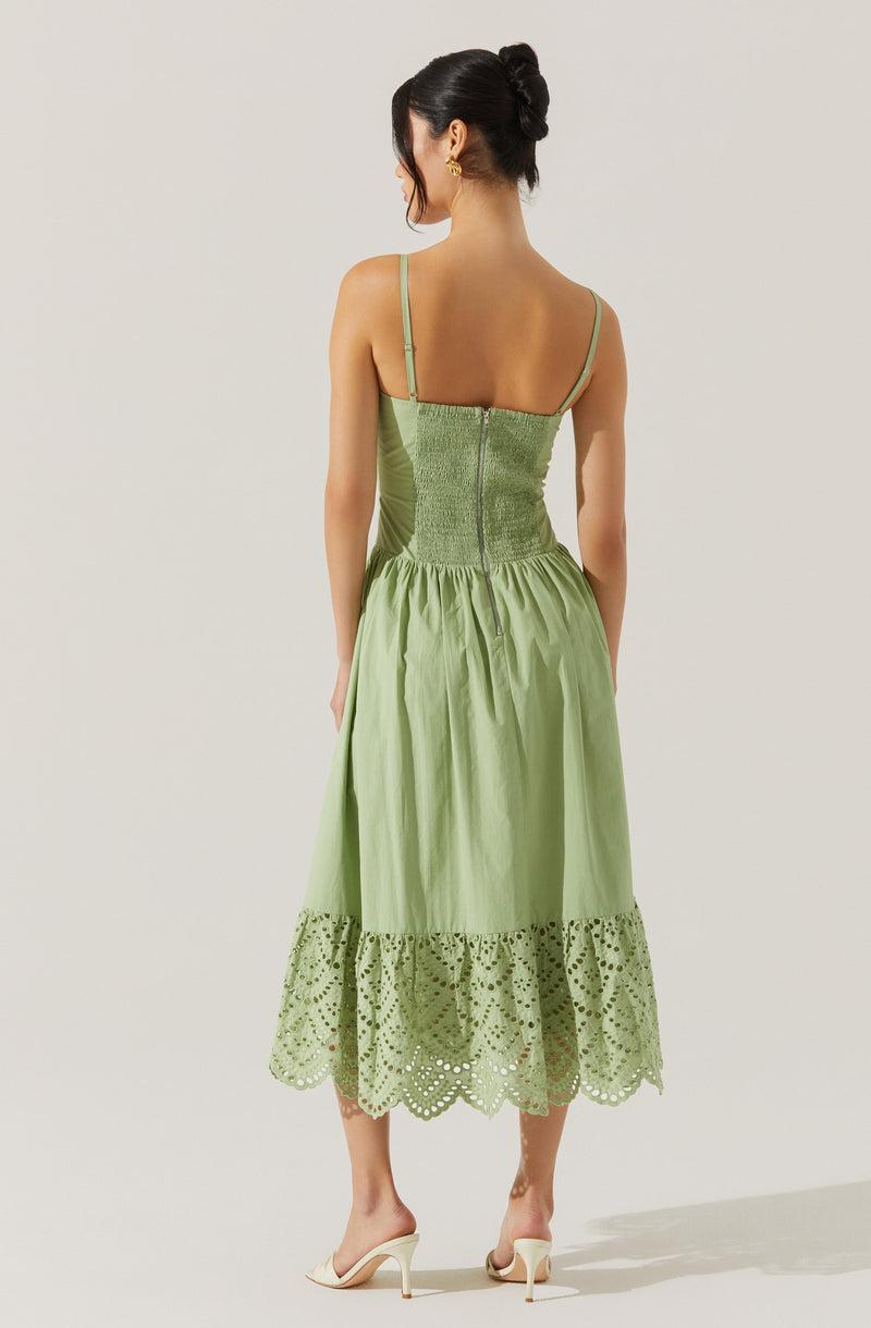 The dress is a sleeveless, light green garment with thin spaghetti straps. The upper part features a fitted, ruched bodice with a visible center zipper at the back. The skirt portion is gathered and has a wide, embroidered hem with eyelet lace detailing, ending mid-calf. The woman is wearing white high-heeled shoes.