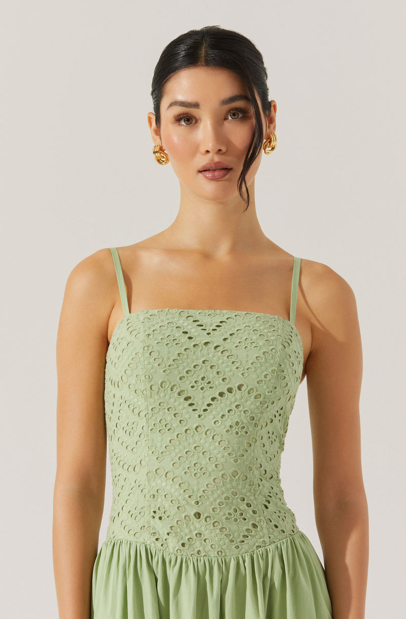 A woman is wearing a light green, sleeveless dress with thin spaghetti straps. The top portion of the dress features a textured, eyelet embroidery pattern with geometric and floral shapes, creating a lace-like appearance. The skirt of the dress is gathered and flows gently from the waist, made of a smooth, lightweight fabric.