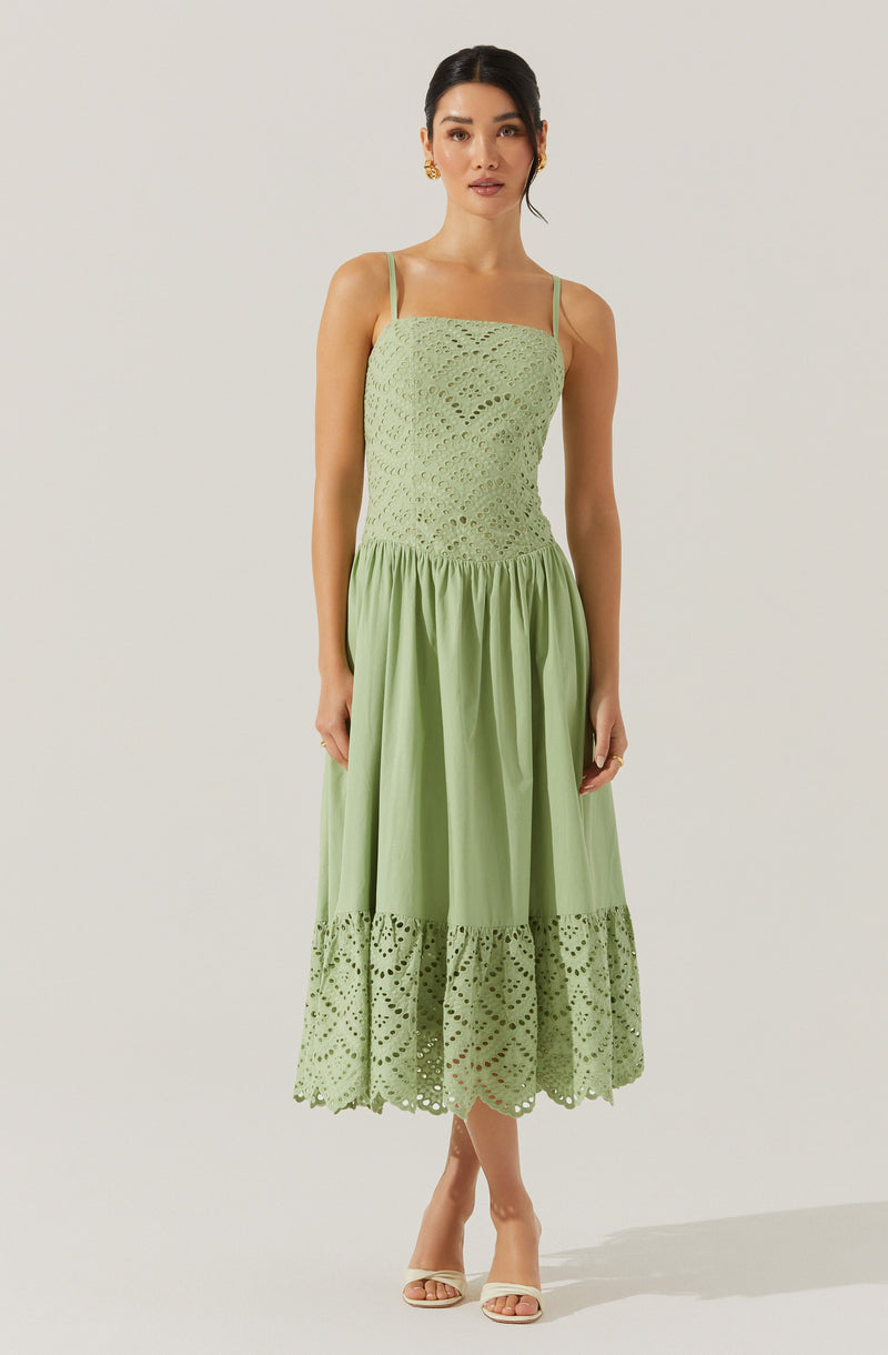 A woman is wearing a light green, sleeveless dress with thin spaghetti straps. The fitted bodice features a delicate, openwork crochet pattern, while the skirt is gathered at the waist with a solid section above a lace-trimmed hem that has a matching crochet pattern. She is also wearing beige open-toe high-heeled sandals and gold earrings.
