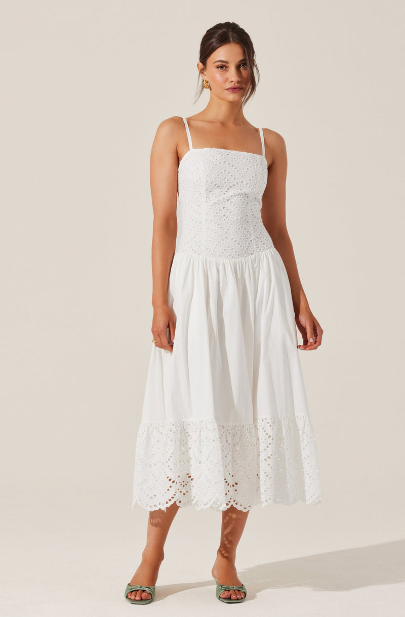 A woman is wearing a white, sleeveless dress with thin spaghetti straps. The dress has eyelet embroidery detail on the bodice and the hem, with a gathered, flowy skirt that reaches mid-calf. She is also wearing green open-toe heels.