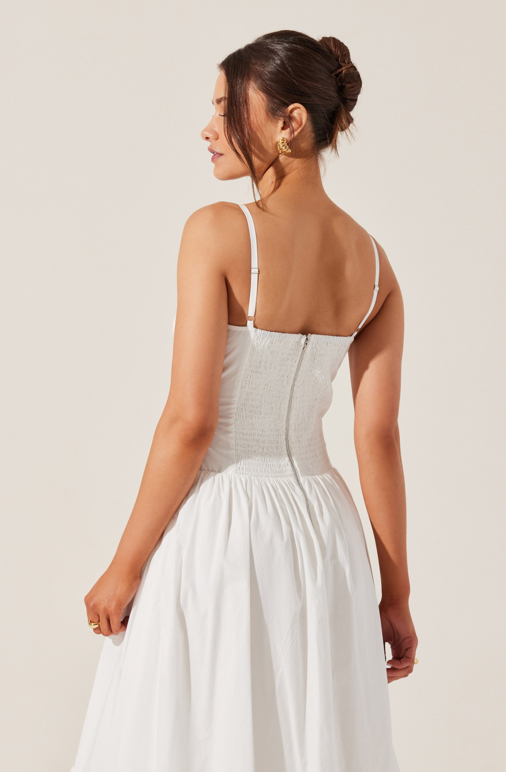 A woman is wearing a white dress with adjustable spaghetti straps. The dress has a fitted, smocked bodice with a zipper at the back, and a full, gathered skirt made of lightweight fabric. She has her hair styled in an elegant updo and is accessorized with gold earrings.