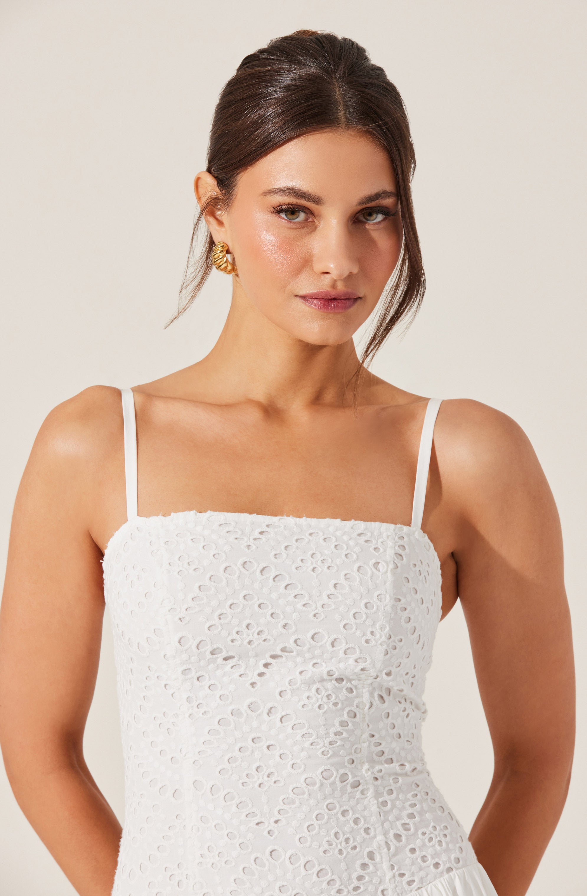 A woman wearing a white sleeveless dress with thin spaghetti straps. The dress has a textured eyelet pattern with circular cutouts. She accessorizes with gold earrings.