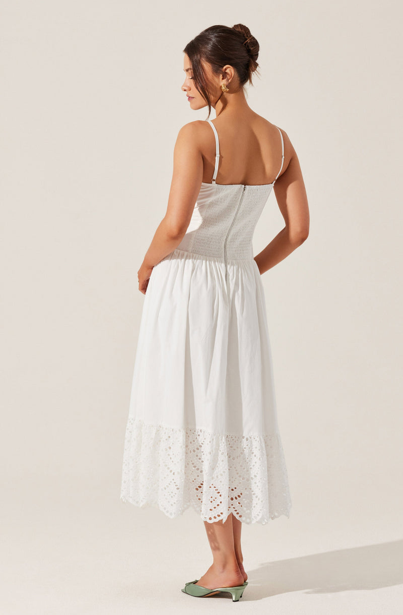 A woman is wearing a white dress with thin spaghetti straps. The top part of the dress is fitted and has a subtle textured pattern, while the skirt is gathered and flows to mid-calf length with eyelet embroidery near the hem. She is also wearing light green heels with a low heel.
