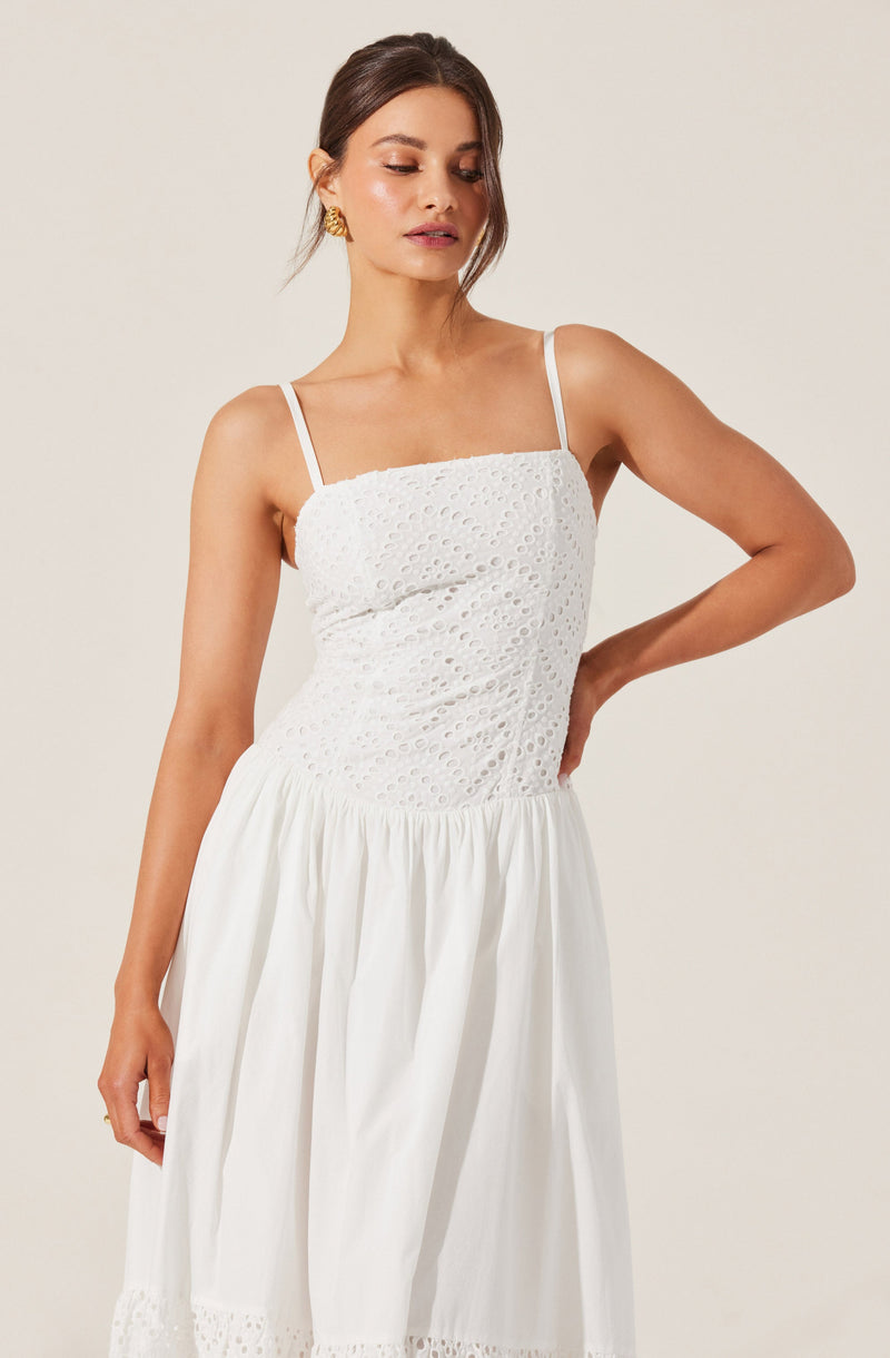 A woman is wearing a white dress with spaghetti straps. The top part of the dress features eyelet embroidery with a floral pattern, while the skirt is gathered and has a simple hem. She has her hair styled in a loose, low ponytail and is accessorized with gold earrings.