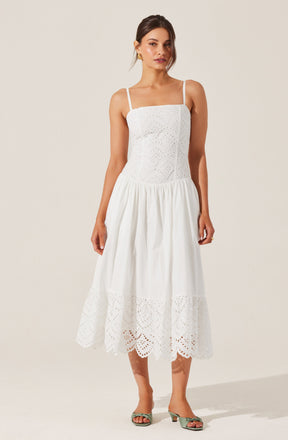 A woman is wearing a white, sleeveless, midi-length dress with spaghetti straps. The dress features eyelet embroidery on the fitted bodice and a gathered, full skirt. The hem of the skirt has a scalloped edge with embroidered cutouts.