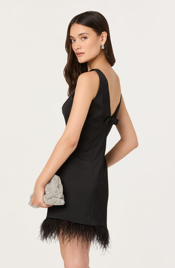 A woman is wearing a sleeveless black dress with wide shoulder straps and a bow detail at the back. The dress features feathers along the hemline. She is holding a silver, sparkly clutch bag and is accessorized with dangling earrings.