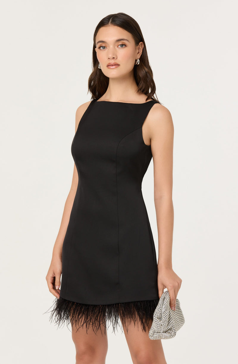 A woman is wearing a sleeveless, fitted black dress with a high neckline. The dress is knee-length and features a feathered trim at the hem. She is holding a small, silver, textured clutch bag in her right hand and has her hair styled in loose waves. She is also wearing earrings.