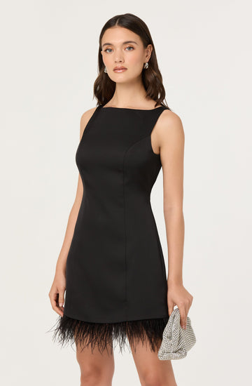 A woman is wearing a sleeveless, fitted black dress with a high neckline. The dress is knee-length and features a feathered trim at the hem. She is holding a small, silver, textured clutch bag in her right hand and has her hair styled in loose waves. She is also wearing earrings.
