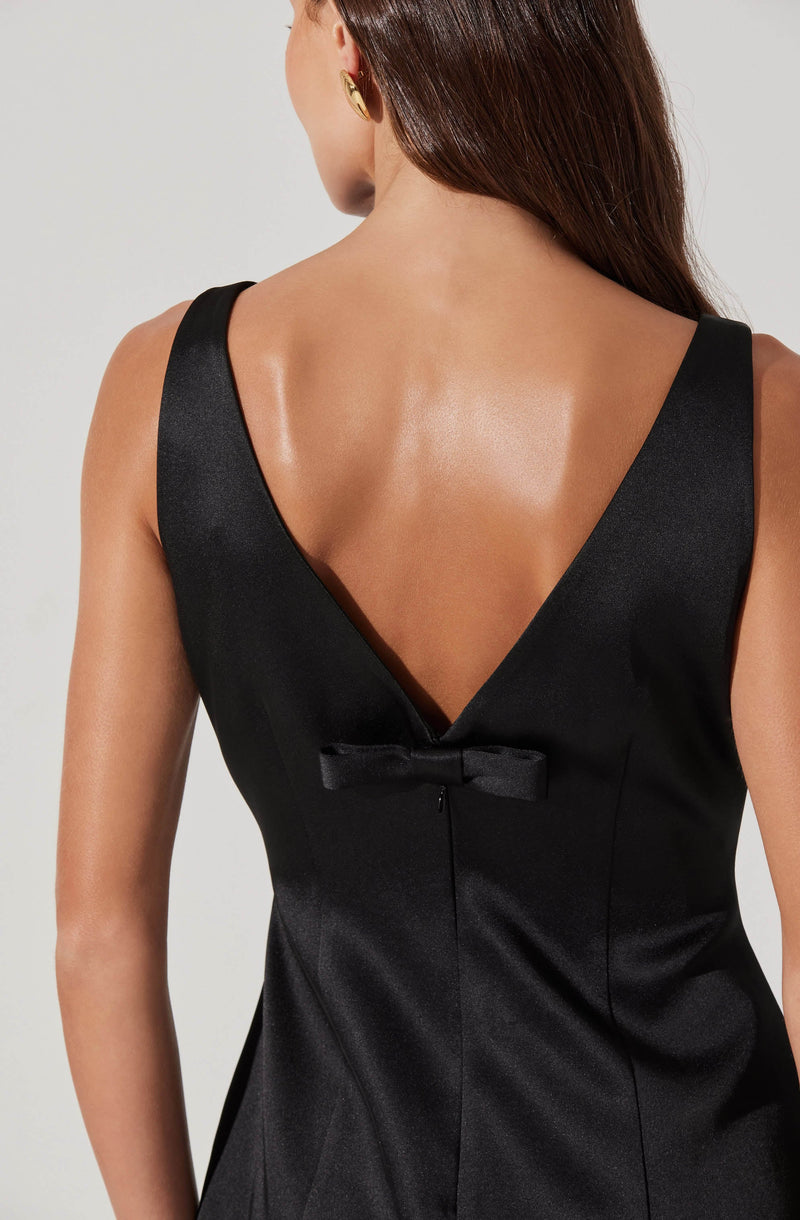 A woman is wearing a sleeveless black dress made from a shiny satin-like fabric. The dress features wide shoulder straps, a deep V-shaped back neckline, and a small decorative bow at the waistline. She has long, dark hair and is accessorized with large, round gold earrings.