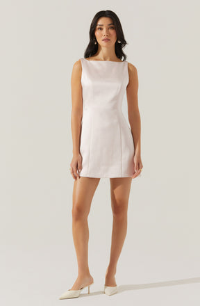 A woman is wearing a sleeveless, ivory-colored, fitted mini dress with a boat neckline and subtle vertical seam details. She pairs the dress with pointed-toe, ivory-colored high heels.