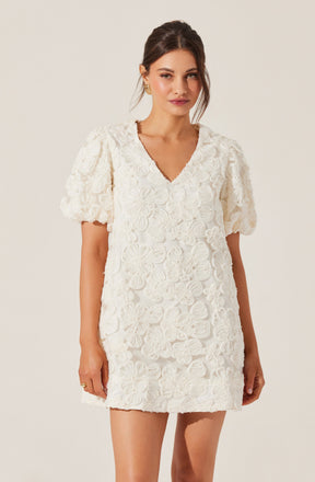 A woman is wearing a white dress with a textured floral pattern. The dress features puffed short sleeves and a V-neckline, with intricate flower motifs covering the entire garment.