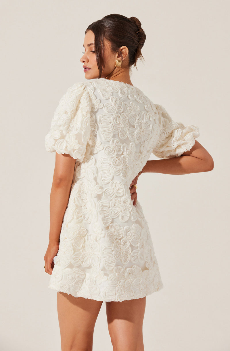 The dress is white with a textured floral pattern. It features puffed short sleeves with ruffle details at the edges. The dress has a fitted waist and a short skirt length.