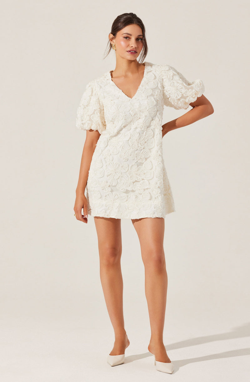 A woman is wearing a short white dress with a floral textured pattern. The dress features puffed sleeves and a V-neckline. She has paired the outfit with white high heels.