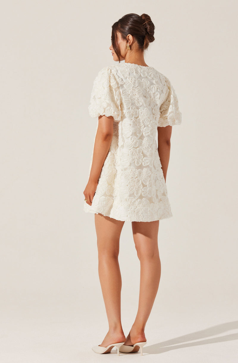 A woman is wearing a white dress with a textured floral pattern. The dress has short, puffed sleeves and a slightly flared hem. She is also wearing white high-heeled shoes.