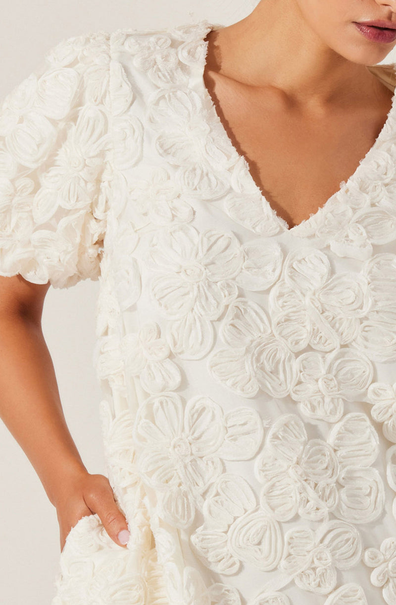 A woman is wearing a white dress with a textured floral pattern. The dress features large, fabric-rolled flower appliqués covering the entire front and short puffed sleeves. It has a deep V-neckline with a frayed edge. The dress appears loose-fitting, with her left hand resting in a side pocket.