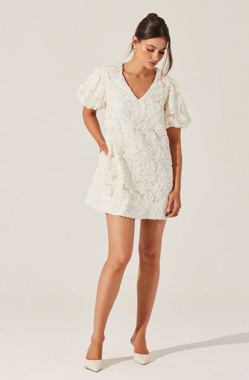 A woman is wearing a short cream-colored dress with textured floral embroidery. The dress has short, puffed sleeves and a V-neckline. She completes her outfit with matching cream-colored high heels.