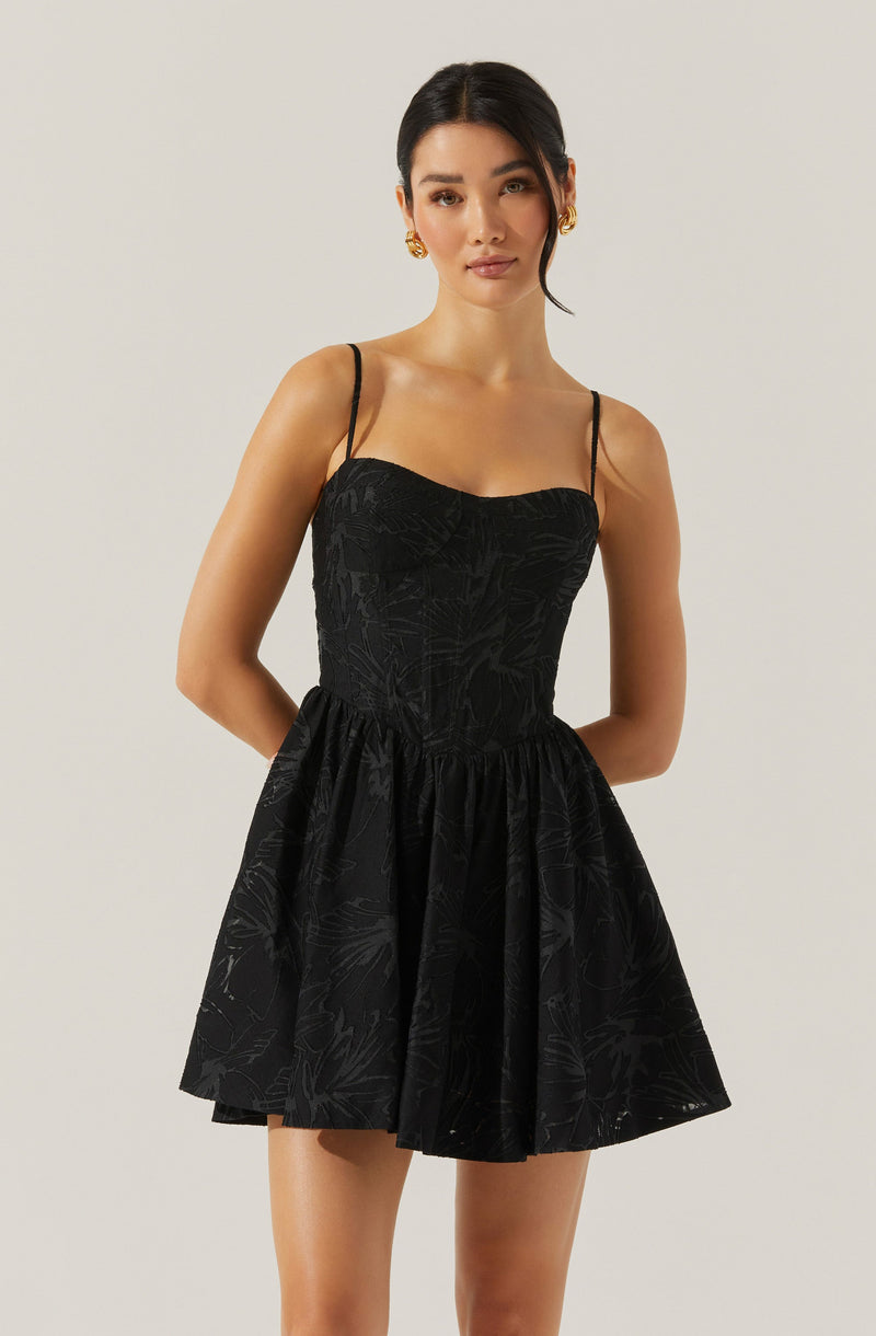A woman is wearing a black dress with thin spaghetti straps. The bodice of the dress is fitted with a subtle textured pattern, and the skirt is gathered and flares out, creating a short, A-line silhouette. The dress features a faint, abstract leaf or floral pattern in a slightly darker shade of black.