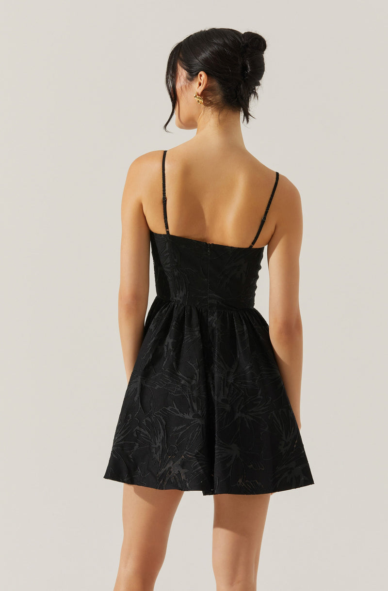 A woman is wearing a black dress with thin spaghetti straps. The dress features a fitted bodice and a flared skirt with a subtle floral pattern. The fabric appears to have a slight sheen and textured design.